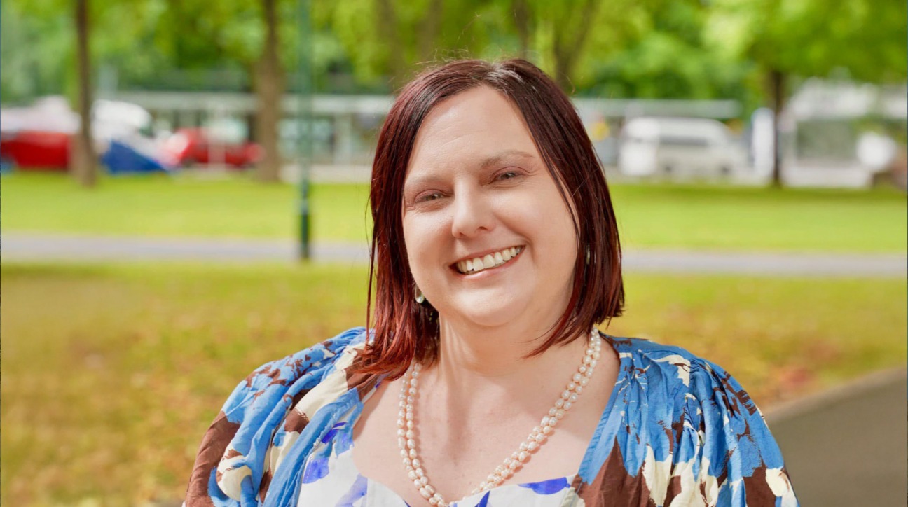 Dr Amy Taylor smiling outdoors, wearing a floral blouse and pearl necklace, with a park setting and blurred trees and vehicles in the background.