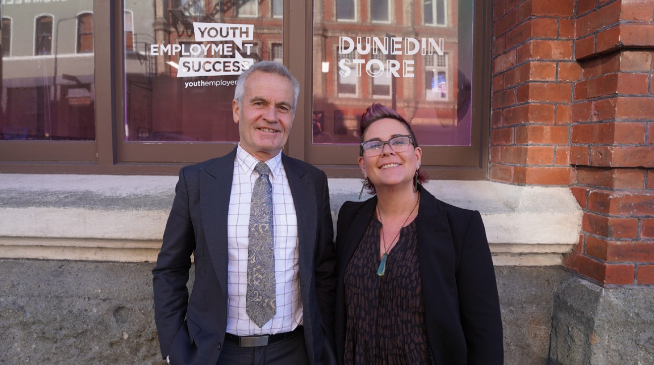 Andrew Simms and Bex Twemlow standing outside the Dunedin Store, smiling in front of a window with “Youth Employment Success” and “Dunedin Store” signage visible.