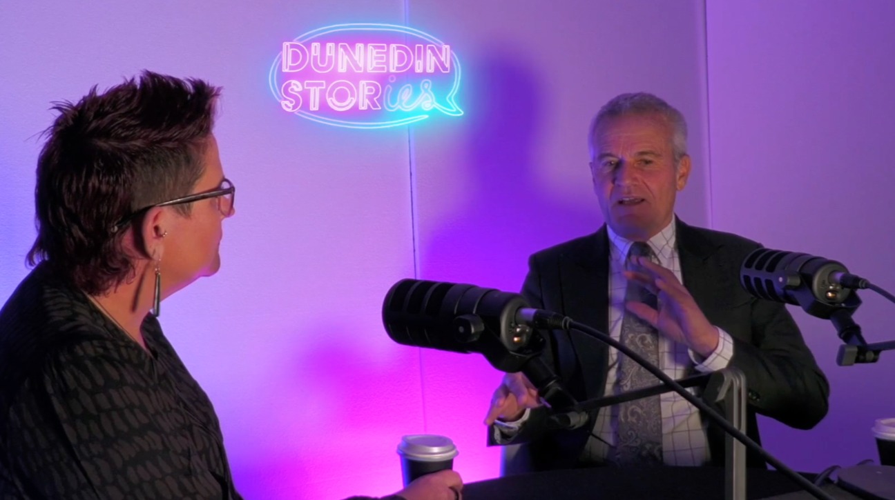 Andrew Simms being interviewed by Bex Twemlow on the Dunedin Stories podcast, seated in a studio with purple lighting and microphones, with the neon Dunedin Stories sign glowing in the background.