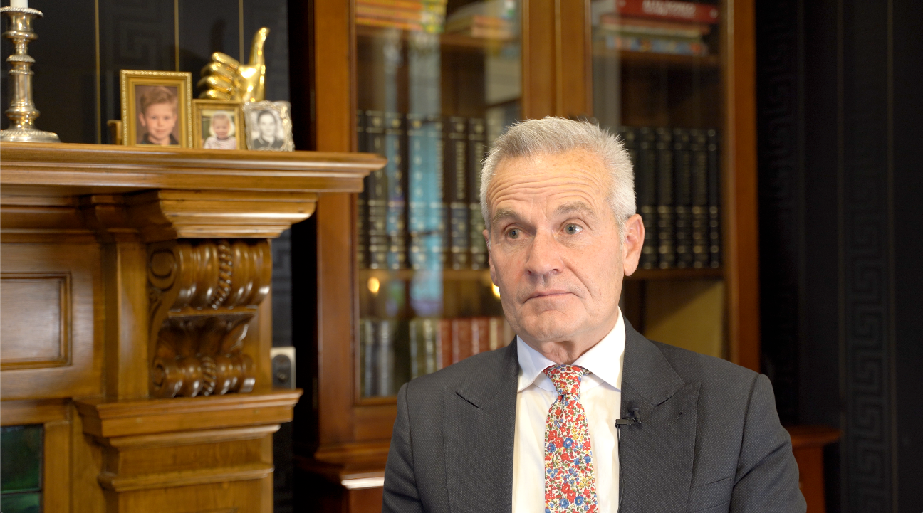 Andrew Simms speaking in a formal setting, wearing a suit and floral tie, seated beside a wooden mantel with family photos and a bookshelf in the background.