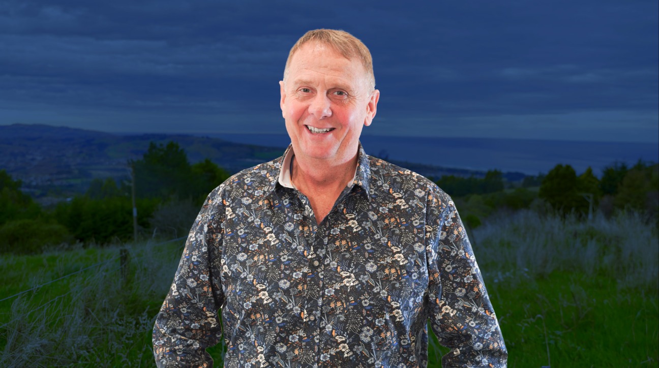 Andrew Sutton standing outdoors in front of a scenic hillside landscape at dusk, wearing a patterned floral shirt and smiling at the camera.