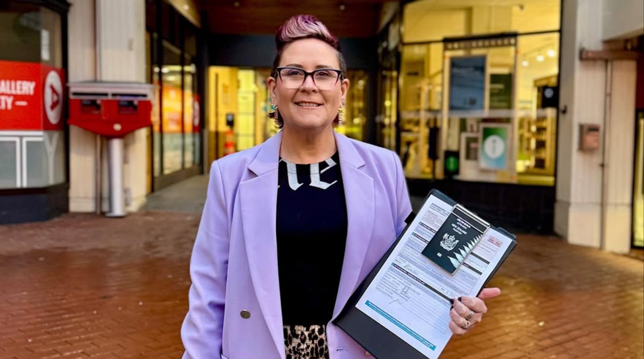 Rebecca (Bex) Twemlow standing outside the Dunedin Electoral Office, smiling and holding her official Council nomination papers and New Zealand passport.