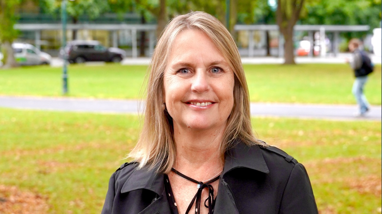 Jo Galer smiling outdoors in a park setting, wearing a black coat, with blurred trees and buildings in the background.