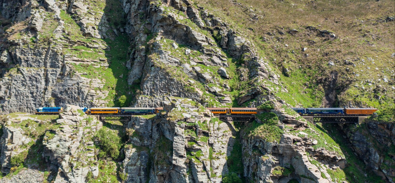  A Dunedin Railways train travelling through the dramatic rock formations of the Taieri Gorge, showcasing steep cliffs, lush greenery, and clear blue skies.
